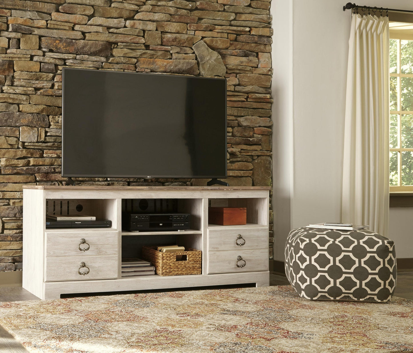 A flat-screen TV sits on the Willowton LG TV Stand w/Fireplace Option; a decorative pouf, stone accent wall, window with curtains, and a patterned rug complete this inviting room.