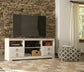 A flat-screen TV sits on the Willowton LG TV Stand w/Fireplace Option; a decorative pouf, stone accent wall, window with curtains, and a patterned rug complete this inviting room.