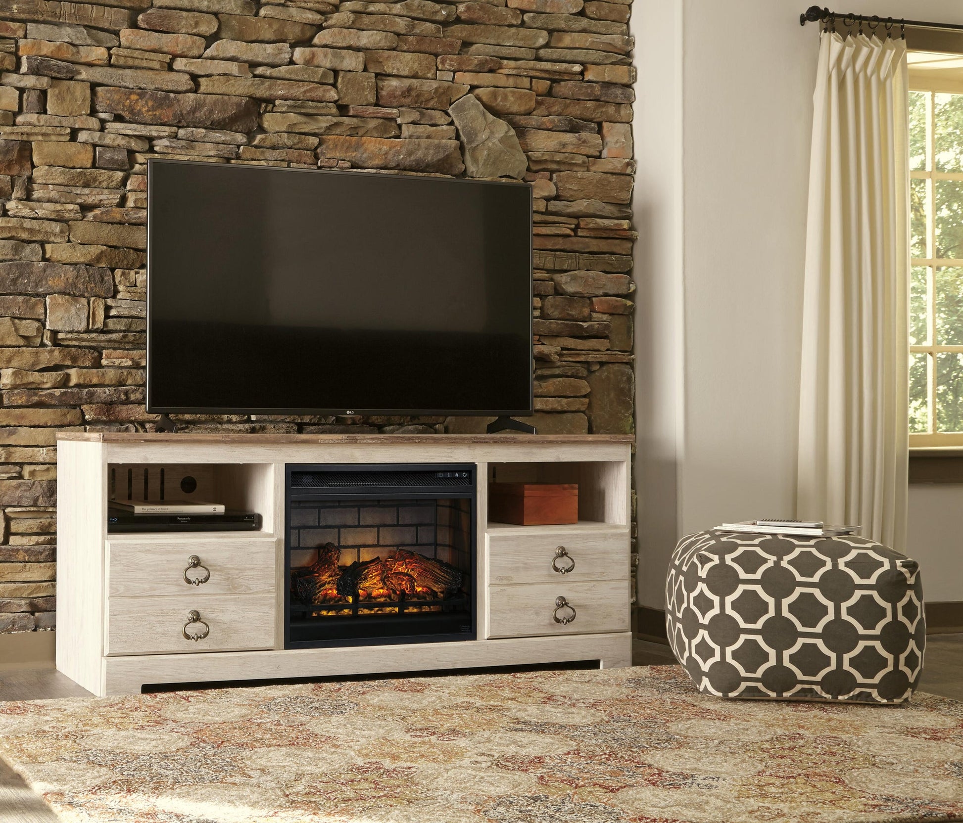 A Willowton LG TV Stand with Fireplace Option holds a flat-screen TV, set against a stone wall, next to a patterned pouf and a window with white curtains.