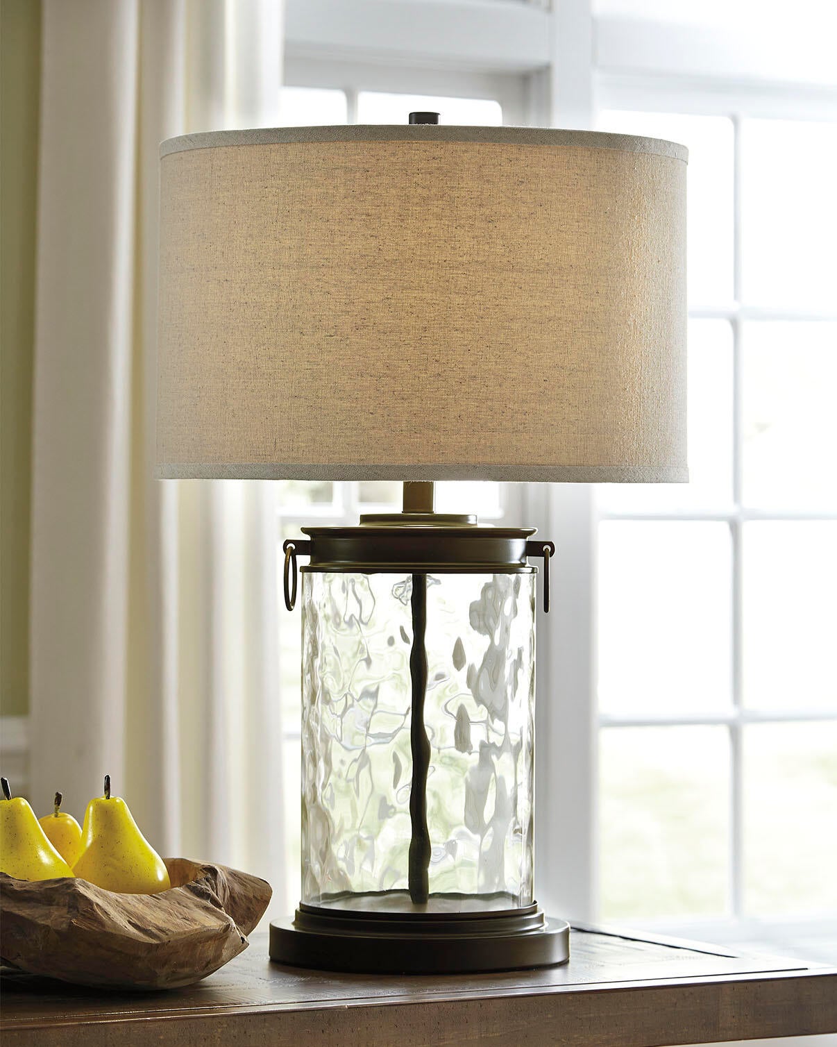 The Tailynn Glass Table Lamp, featuring a textured cylindrical base and beige fabric shade, sits on a wooden surface near a window beside a wooden bowl filled with yellow pears.