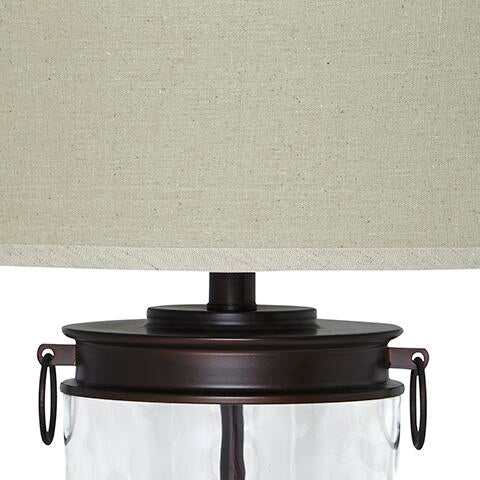 Close-up of the Tailynn Glass Table Lamp (1/CN), featuring a beige fabric drum shade, dark metal neck with side handles, and a clear wave-patterned glass base.