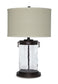 The Tailynn Glass Table Lamp (1/CN) features a clear textured cylindrical glass base, dark metal accents, and a large beige drum shade, providing stylish illumination.
