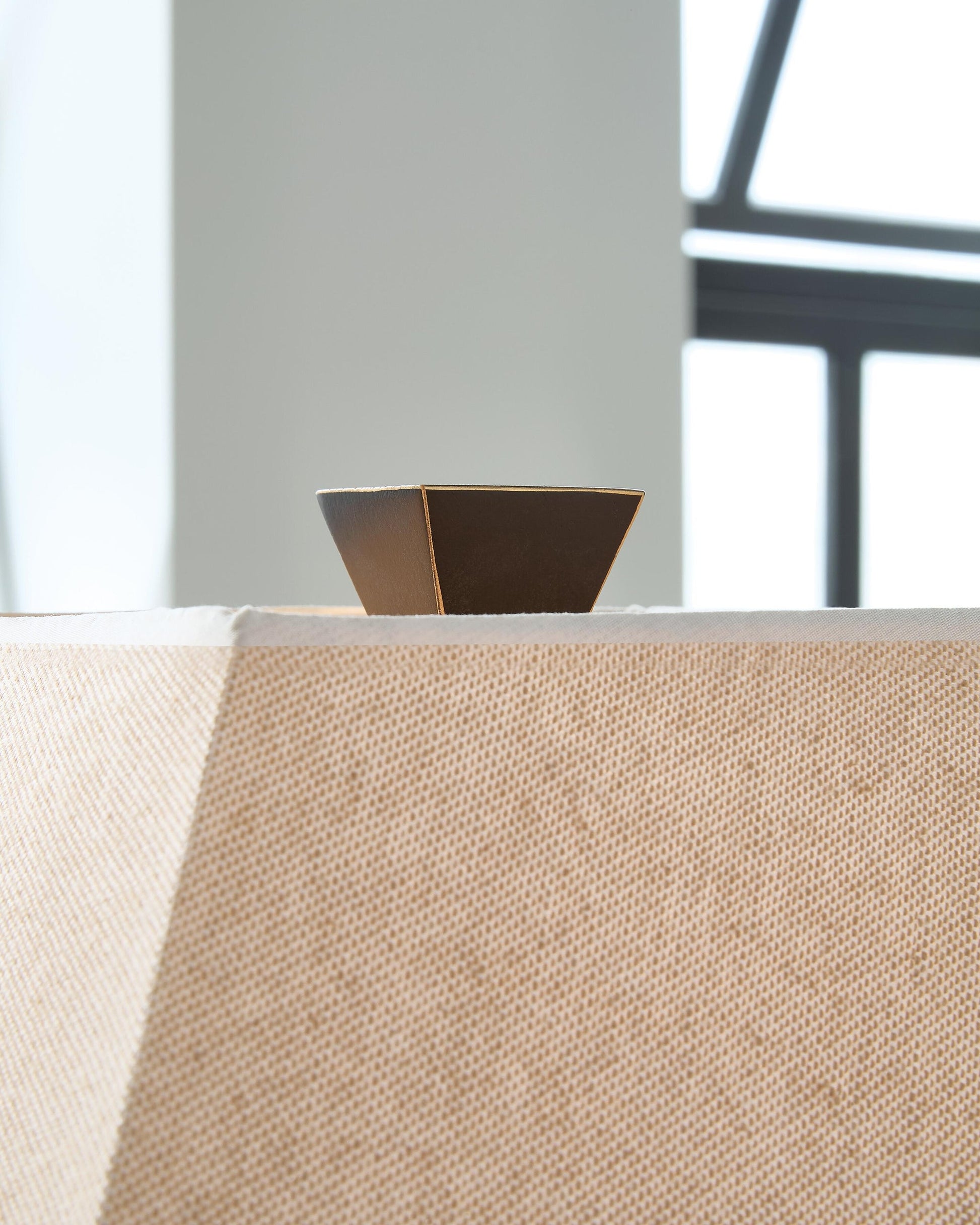 Close-up of the brown, trapezoidal finial atop the beige, linen-look shade of the Deidra Metal Table Lamp (2/CN), with a window in the background.