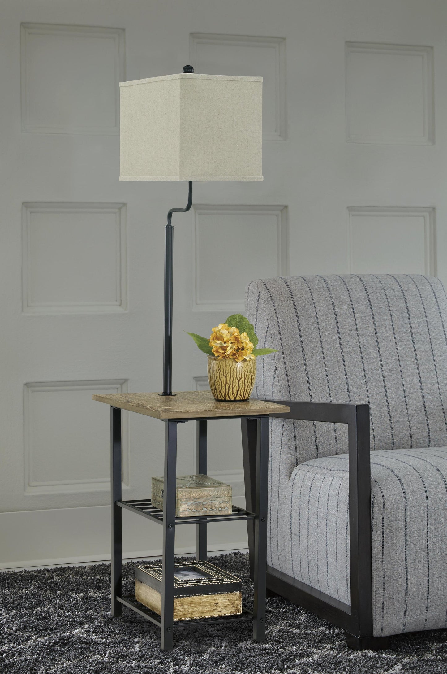A square side table with a space-saving Shianne Metal Tray Lamp, a potted yellow flower, and two books stands next to a gray striped armchair on a textured dark rug.