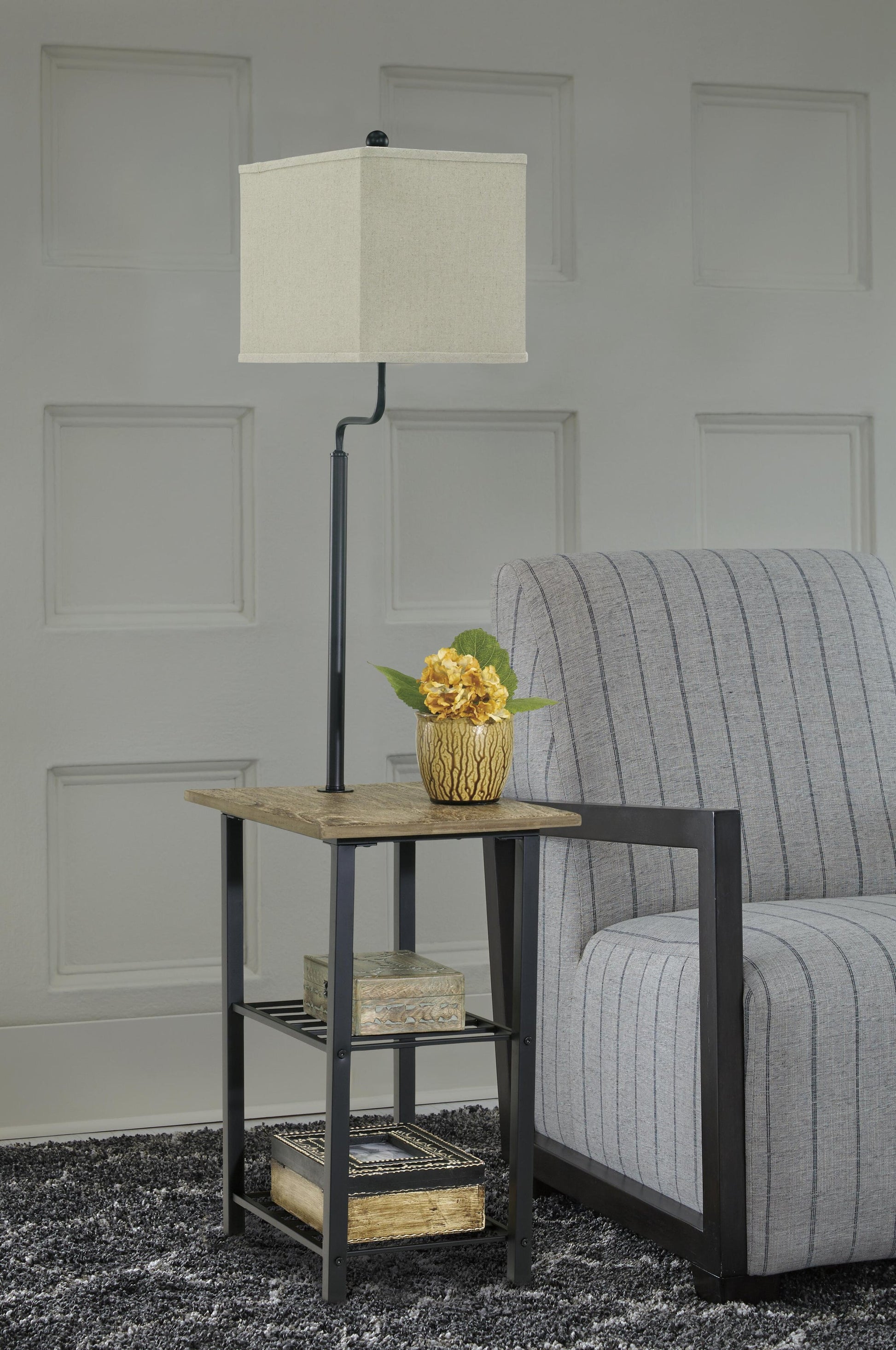 A square side table with a space-saving Shianne Metal Tray Lamp, a potted yellow flower, and two books stands next to a gray striped armchair on a textured dark rug.