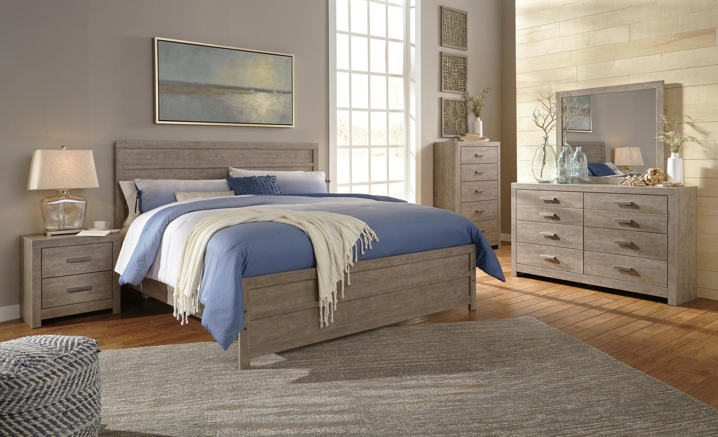 A modern bedroom with a wooden bed, blue bedding, nightstand, lamp, Culverbach Six Drawer Dresser with mirror, tall chest, large window, wall art, and a rug on hardwood floor—perfect for stylish bedroom storage furniture.